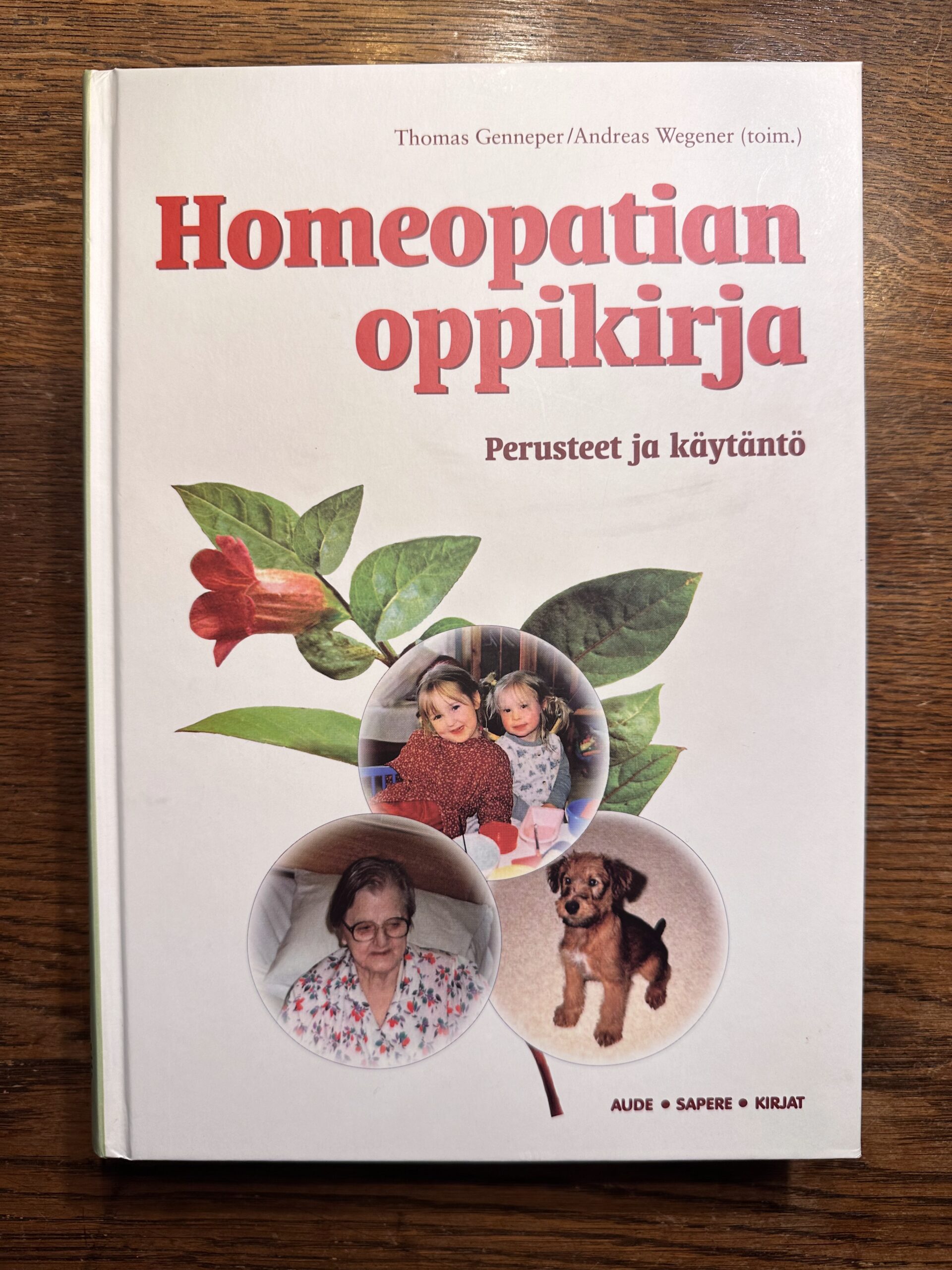 Kuvassa kirja jonka nimi on: Homeopatian oppikirja. Siinä on kuva punaisesta kukasta ja kolme kuplaa joissa yhdessä nainen, toisessa kaksi lasta ja kolmannessa koira.